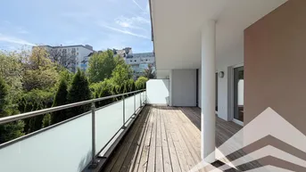 Große Terrasse mit Holzboden und Glasgeländer, umgeben von Bäumen und Gebäuden unter blauem Himmel.