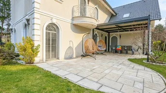 Große gepflasterte Terrasse mit Hängesesseln und Überdachung, umgeben von Rasen und Garten.