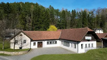Gebäude mit rotem Ziegeldach und weißer Fassade, umgeben von grünen Hügeln und Wald.