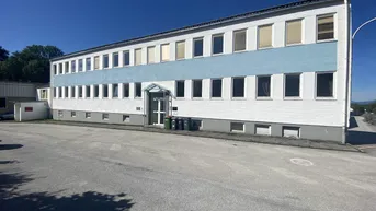 Gebäude mit weißer Fassade und blauem Band, umgeben von einem Parkplatz unter blauem Himmel.