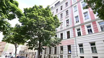 Klassisches Mehrfamilienhaus mit rosafarbener Fassade, umgeben von Bäumen und parkenden Autos auf der Straße.