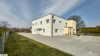 Modernes weißes Gebäude mit Flachdach, gepflasterter Auffahrt und gepflegter Rasenfläche in ländlicher Umgebung.
