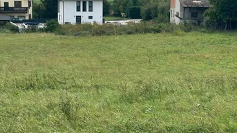 Blick über eine grüne Wiese auf ein modernes Haus und ein älteres Gebäude mit Bergkulisse im Hintergrund.