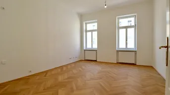 Helles Zimmer mit Fischgrätparkett, hohen Decken und zwei großen Fenstern.