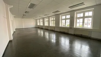 Geräumiger, heller Raum mit zahlreichen Fenstern, dunklem Boden und praktischer Rasterdecke.