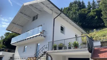 Modernes weißes Haus mit Balkon und Terrasse, eingebettet in eine grüne Hügellandschaft.
