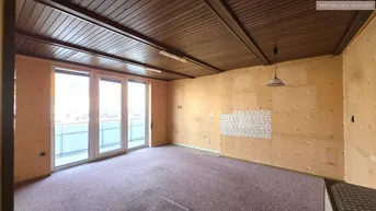 Geräumiger Innenraum mit Teppichboden, Holzdecke und Zugang zu einem Balkon, renovierungsbedürftig.