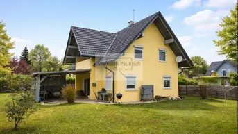 Gelbes Einfamilienhaus mit großem Garten und überdachtem Stellplatz unter blauem Himmel.