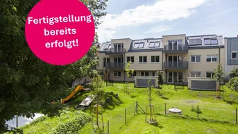Modernes Mehrfamilienhaus mit Balkonen, gepflegtem Garten und Kinderspielplatz unter blauem Himmel.