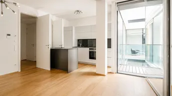 Heller offener Wohnbereich mit moderner Küche, Holzboden und Zugang zu einem verglasten Balkon.