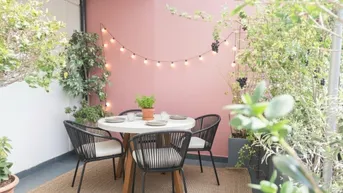Gemütliche Terrasse mit rosafarbener Wand, Lichterkette und Pflanzen für entspannte Abende.