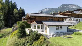 Traditionelles Holzhaus in alpiner Umgebung mit grünen Hügeln und bewaldeten Bergen im Hintergrund.