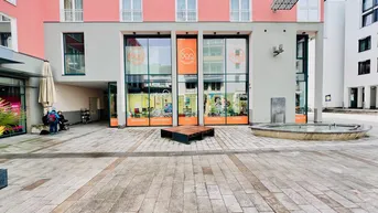 Moderne Geschäftsfassade mit großen Schaufenstern und einem Brunnen auf dem gepflasterten Vorplatz in urbaner Umgebung.