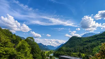 Panoramablick auf grüne Berge, ein Tal und einen Fluss unter weitem Himmel.