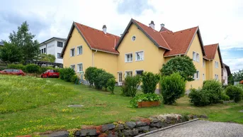 Gelbes Mehrfamilienhaus mit rotem Ziegeldach und gepflegter Grünfläche in ländlicher Umgebung.