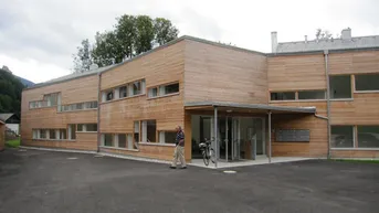 Moderne Wohnanlage mit Holzfassade und großzügigem Eingangsbereich, umgeben von grüner Landschaft.