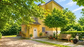 Charmantes gelbes Haus mit gepflegtem Vorgarten, umgeben von üppigem Grün und Bäumen.