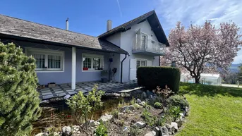 Hausfassade mit gepflegtem Garten, Zierteich und Terrasse, umgeben von blühenden Bäumen unter blauem Himmel.