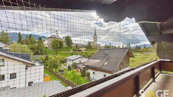 Blick vom Balkon über ein Dorf mit Kirche und umliegender Berglandschaft.
