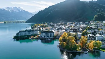 Panoramablick auf eine malerische Stadt am Seeufer, umgeben von majestätischen Bergen und herbstlicher Natur.