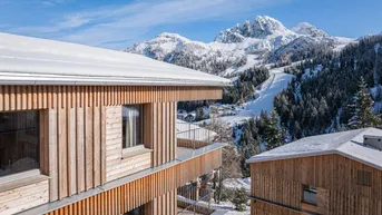 Moderne Holzgebäude mit Balkonen vor einer verschneiten Berglandschaft und Skipisten.