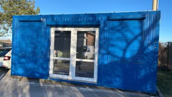 Blauer Containerbau mit Doppeltür und Rollläden, auf einem gepflasterten Bereich stehend.
