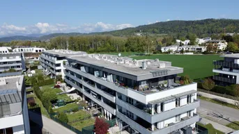 Luftaufnahme moderner Wohngebäude mit Flachdächern, umgeben von Grünflächen und Bergen im Hintergrund.