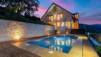 Haus mit beleuchtetem Pool und Holzdeck bei Sonnenuntergang, umgeben von grüner Landschaft.