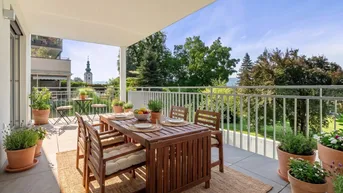 Möblierter Balkon mit Holzesstisch, Pflanzen und weitem Blick ins Grüne mit Kirchturm.