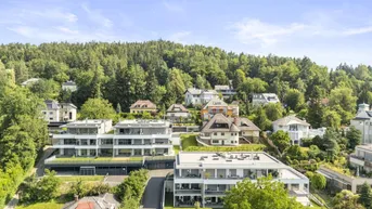 Luftaufnahme einer modernen Wohnsiedlung mit mehreren Gebäuden, umgeben von viel Grün und Hügellandschaft.