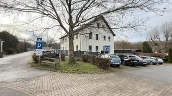 Weißes Mehrfamilienhaus mit Parkplätzen und Bäumen an einer Straße gelegen.