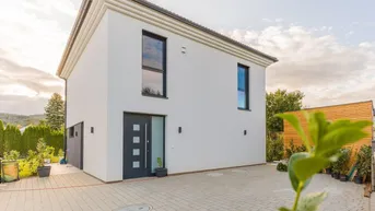 Modernes Einfamilienhaus mit weißer Fassade, dunklen Fenstern und gepflasterter Auffahrt.