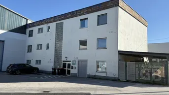Gebäudeaußenansicht mit weißer Fassade, mehreren Fenstern und einem Parkplatzbereich vor dem Eingang.