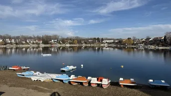 Blick auf einen See mit bunten Tretbooten am Ufer und Häusern im Hintergrund unter blauem Himmel.