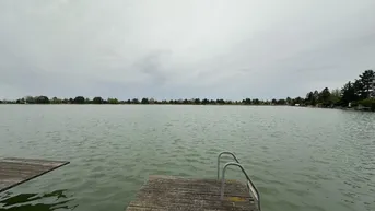 Weitläufiger Blick über einen ruhigen See mit einem Holzsteg im Vordergrund.