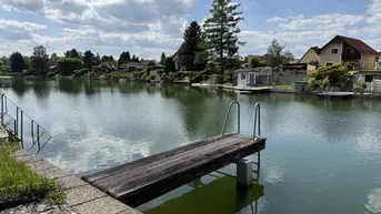 Ruhiger See mit Holzsteg und umliegenden Häusern unter blauem Himmel.