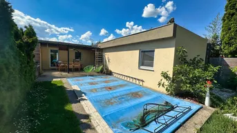 Außenbereich mit überdachtem Pool, Rasenfläche und einer möblierten Terrasse unter blauem Himmel.