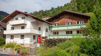 Ansicht des Gebäudes mit traditioneller Architektur, umgeben von grüner Vegetation und einem Berg im Hintergrund.