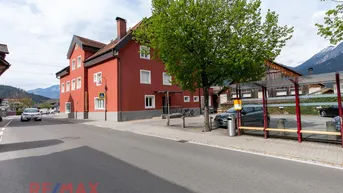 Rotes Gebäude mit traditioneller Architektur an einer Straße, umgeben von Bäumen und Bergen.