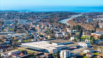 Weitläufige Luftaufnahme einer Stadtlandschaft mit Wohngebieten, Industriegebäuden, einem Fluss und einem See im Hintergrund.