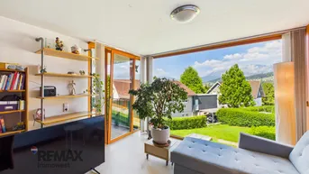 Helles Wohnzimmer mit großen Fenstern, Blick ins Grüne und auf die Berge, ausgestattet mit Sofa und TV.