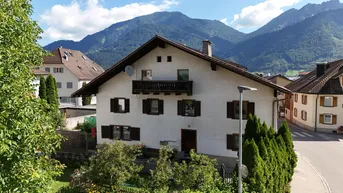 Traditionelles Haus mit Satteldach und Holzbalkon vor einer beeindruckenden Bergkulisse.