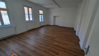 Geräumiger, leerer Raum mit hellem Laminatboden und mehreren Fenstern, die viel Tageslicht hereinlassen.