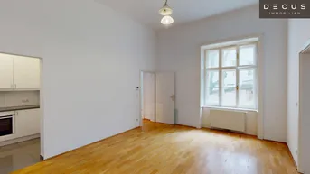 Helles Wohnzimmer mit Parkettboden, hohen Decken und Zugang zu einer separaten Küche.