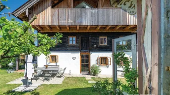 Traditionelles Bauernhaus mit weißer Fassade, Holzbalkonen und einem gemütlichen Gartenbereich mit Sitzgelegenheiten.
