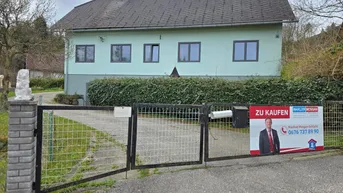 Vorderansicht des Hauses mit hellgrüner Fassade, dunklem Dach und einem Zaun mit Werbeschild.