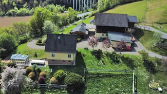 Luftaufnahme eines ländlichen Anwesens mit zwei Gebäuden, einem Gewächshaus und grünen Hügeln im Hintergrund.