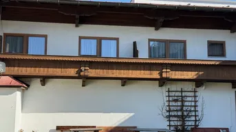 Rückseite eines Gebäudes mit traditionellen Holzbalkonen und Sitzgelegenheiten im Freien vor einer grünen Wiese.