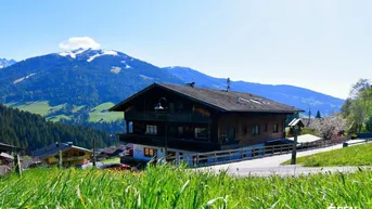 Traditionelles Holzhaus in alpiner Landschaft mit schneebedeckten Bergen im Hintergrund unter blauem Himmel.