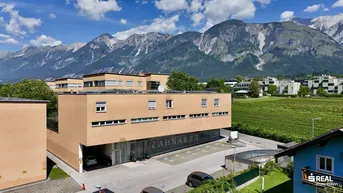 Gebäudeansicht mit Zahnarztpraxis, Parkplätzen und beeindruckendem Alpenpanorama im Hintergrund.